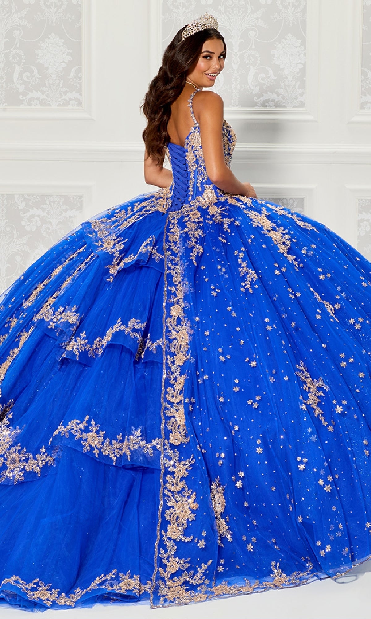 A woman in the Quinceanera Dress PR30087 By Princesa, featuring royal blue fabric and gold embroidery, stands before a white ornate wall with a tiara, looking over her shoulder and smiling.